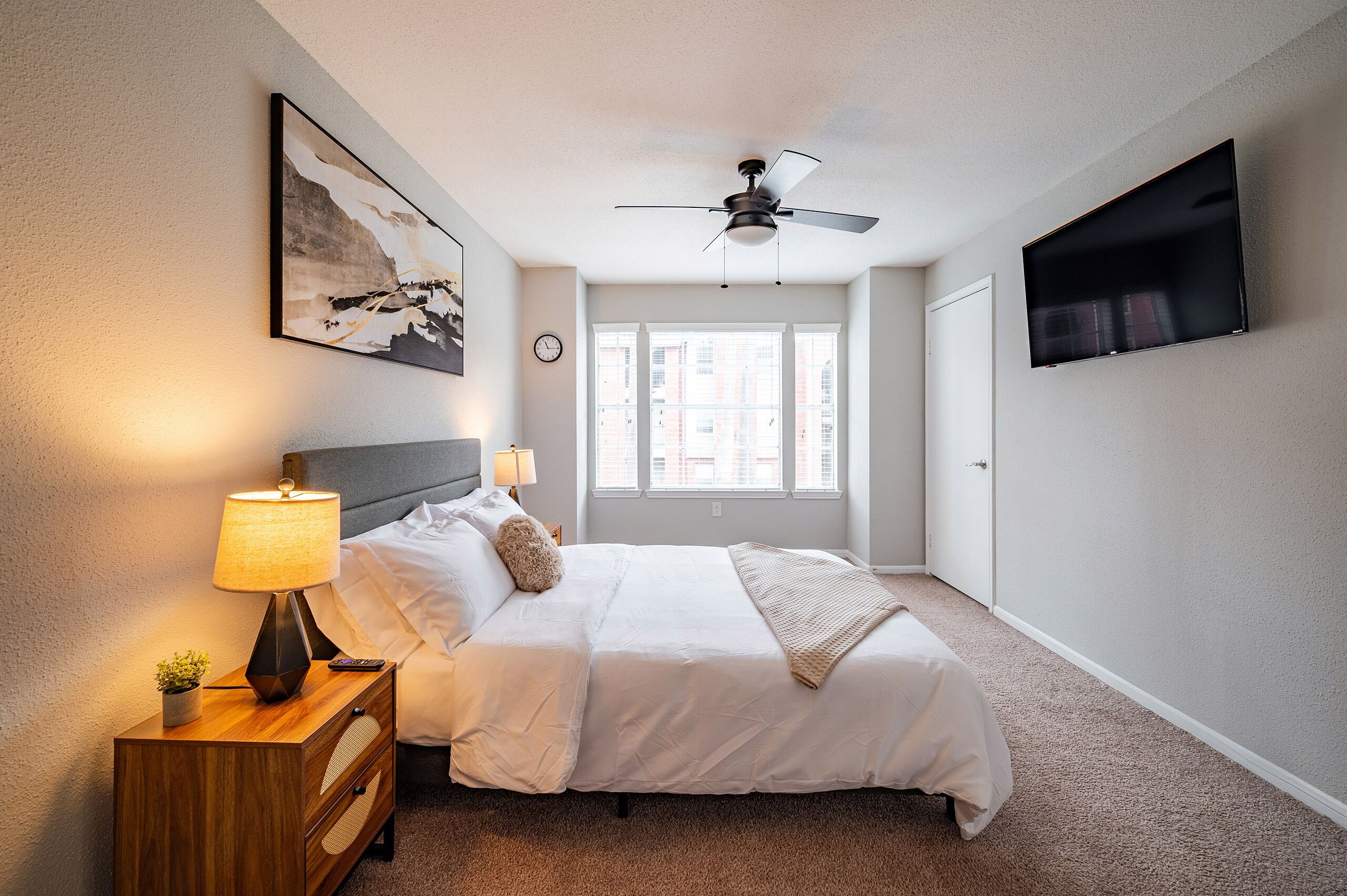 Ein Hotelzimmer mit Bett, Deckenventilator, an der Wand befestigtem Fernseher und einem Fenster mit Blick auf die Stadt.