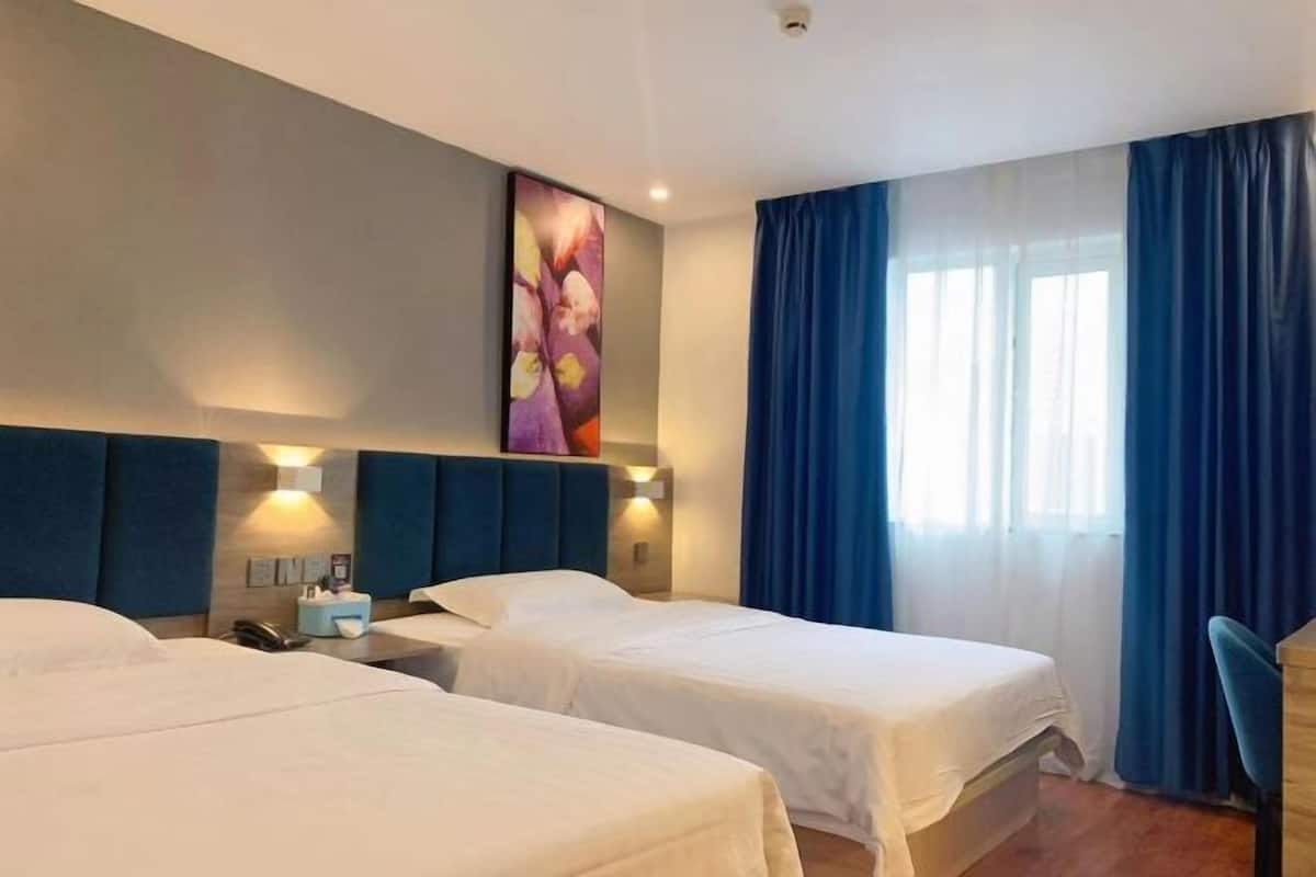 A hotel room with two beds, a large window with blue curtains, a wall-mounted artwork, and a desk with a chair.