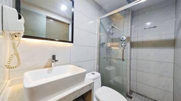 Un baño con un lavamanos blanco, un inodoro, una ducha con mampara de vidrio y un teléfono instalado en la pared.