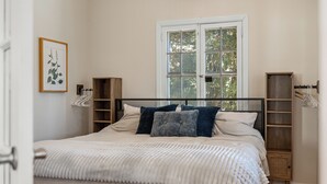 A neatly made bed with a white comforter and pillows, a wooden headboard, a window with a view of trees, and a small wooden cabinet with hanging clothes. - Elevated Potomac River Retreat - King Suite - Riverfront - Large Screened Porch (Sharpsburg)