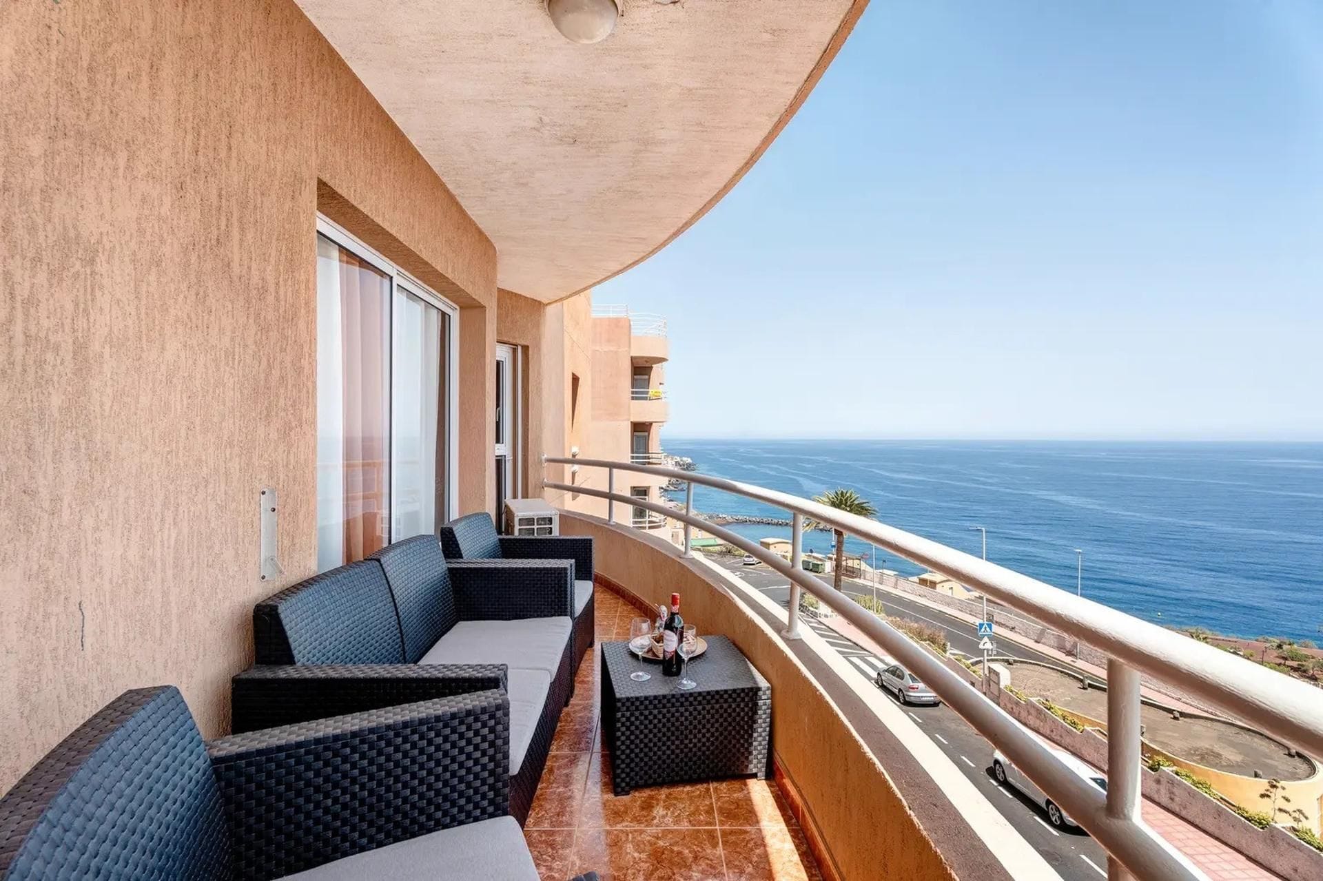 A balcony with outdoor seating, a view of the ocean, and a building with a beige facade.