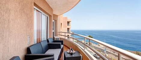 A balcony with outdoor seating, a view of the ocean, and a building with a beige facade.