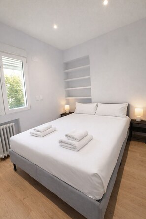 Un dormitorio moderno con una cama grande, dos lámparas de noche, un radiador y una ventana con persianas.