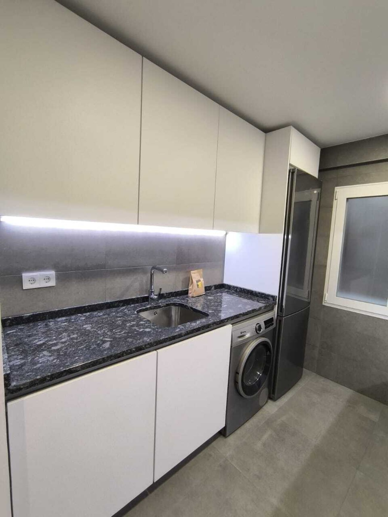 A modern kitchen with white cabinets, a black granite countertop, a stainless steel sink, a washing machine, and a refrigerator.