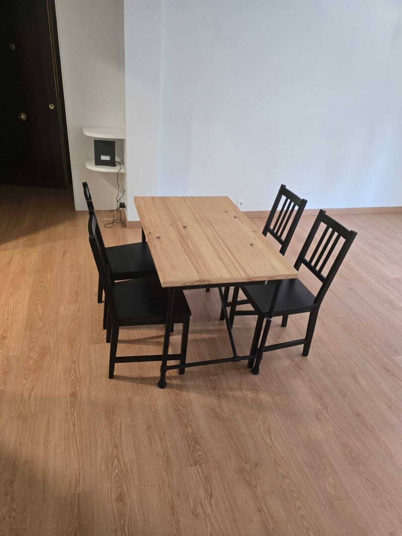 A rectangular wooden table with four black chairs, a white wall, and a wooden floor.