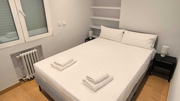 A bedroom with a bed, a window, a radiator, and a nightstand with a lamp.