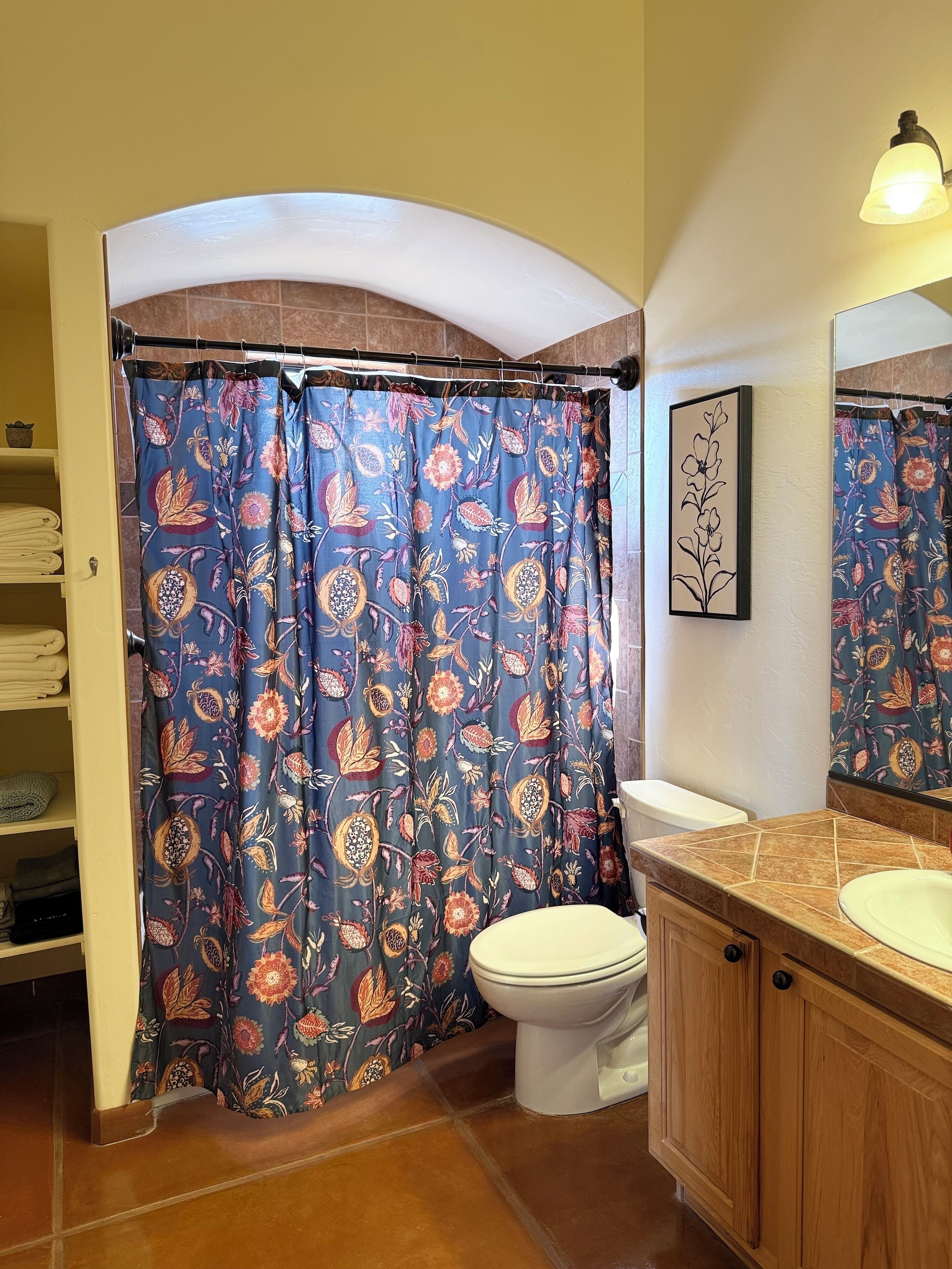 Un baño con cortina de ducha estampada con motivos florales, un inodoro, un lavamanos con espejo y un estante con toallas dobladas.