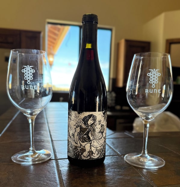 Una botella de vino con una etiqueta detallada, dos copas de vino vacías y un televisor al fondo.