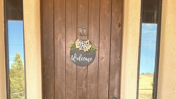 Una puerta de madera con un cartel que dice 'Welcome' encima, flanqueada por dos ventanas y un felpudo negro con la palabra 'welcome'.