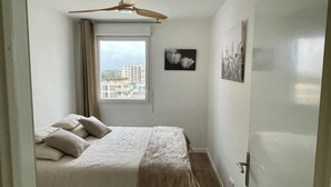 A bedroom with a bed, a ceiling fan, a window with a view of buildings, and two wall-mounted pictures.