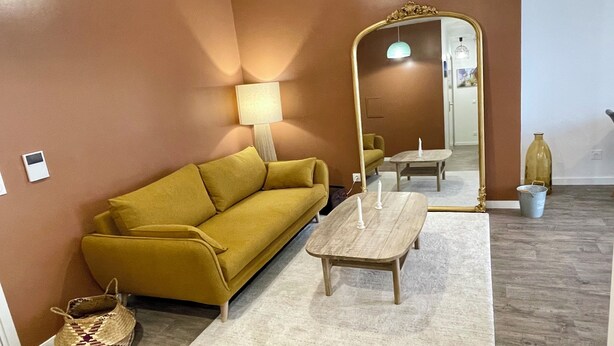 A room with a mustard yellow sofa, a wooden coffee table, a lamp, a mirror, a small table with a candle, a wicker basket, and a metal bucket.