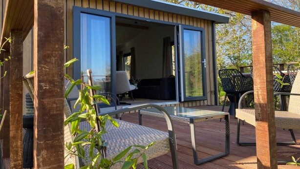 A modern outdoor deck with a glass door leading to a room, surrounded by wooden beams and greenery. - Luxury holiday cottage, stunning views, and hot tub. (Pingjum)
