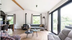 A modern living room with a sofa, two armchairs, a coffee table, and a dining area with chairs and a table. There are large windows with curtains, a ceiling light, and a view of greenery outside. - Luxury holiday cottage, stunning views, and hot tub. (Pingjum)