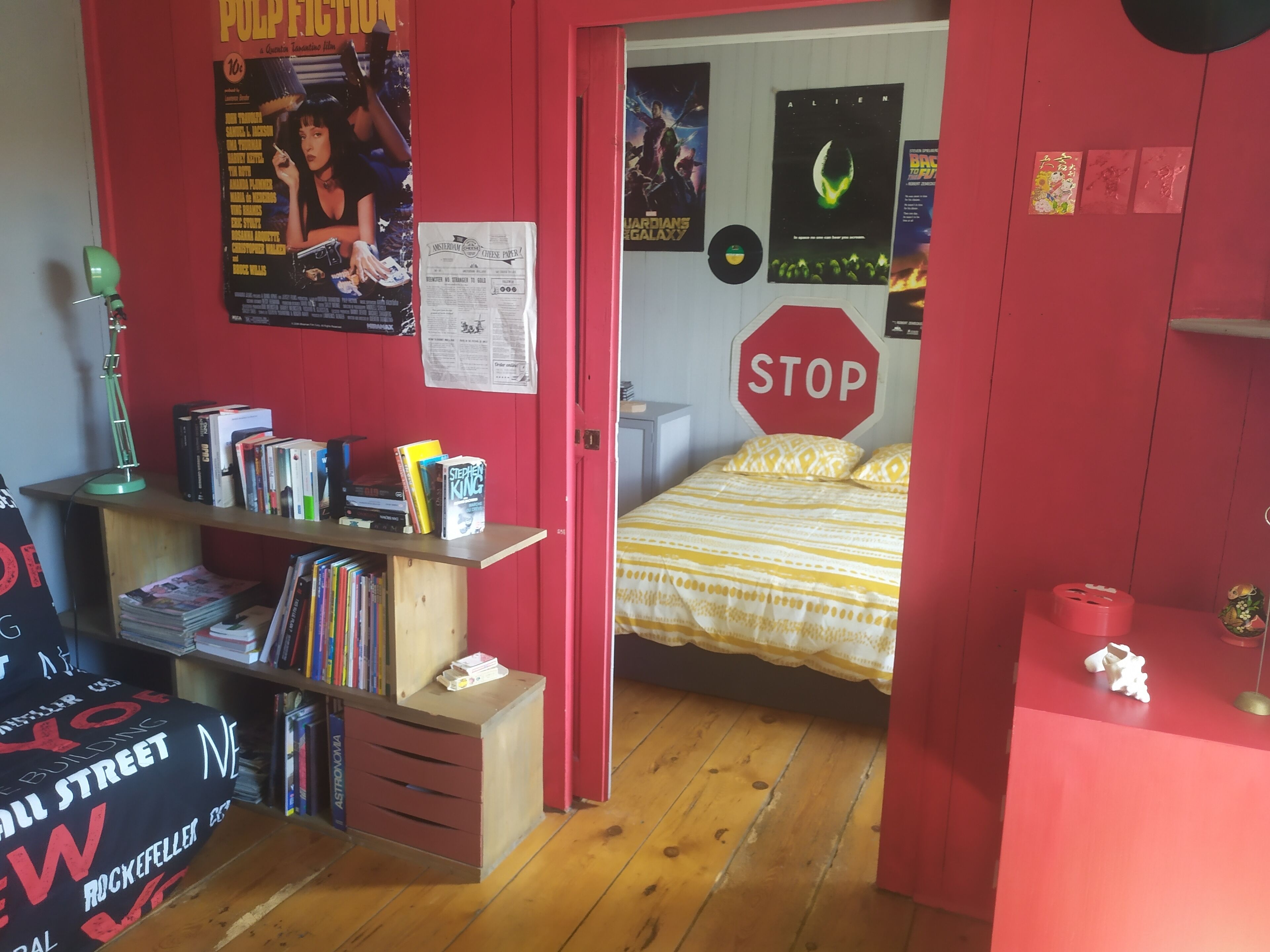 Una stanza con pavimento in legno, un letto con copriletto a righe, una libreria piena di libri, una scrivania con una lampada e un poster appeso al muro.