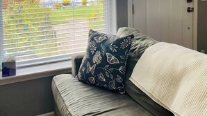 A room with a grey sofa, a window with blinds, a door with a white curtain, and a view of greenery outside.
