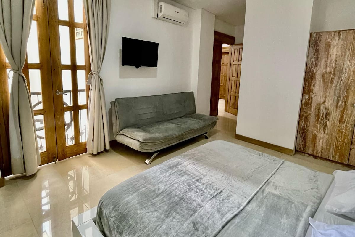 A bedroom with a bed, a sofa, a TV, and an air conditioning unit.