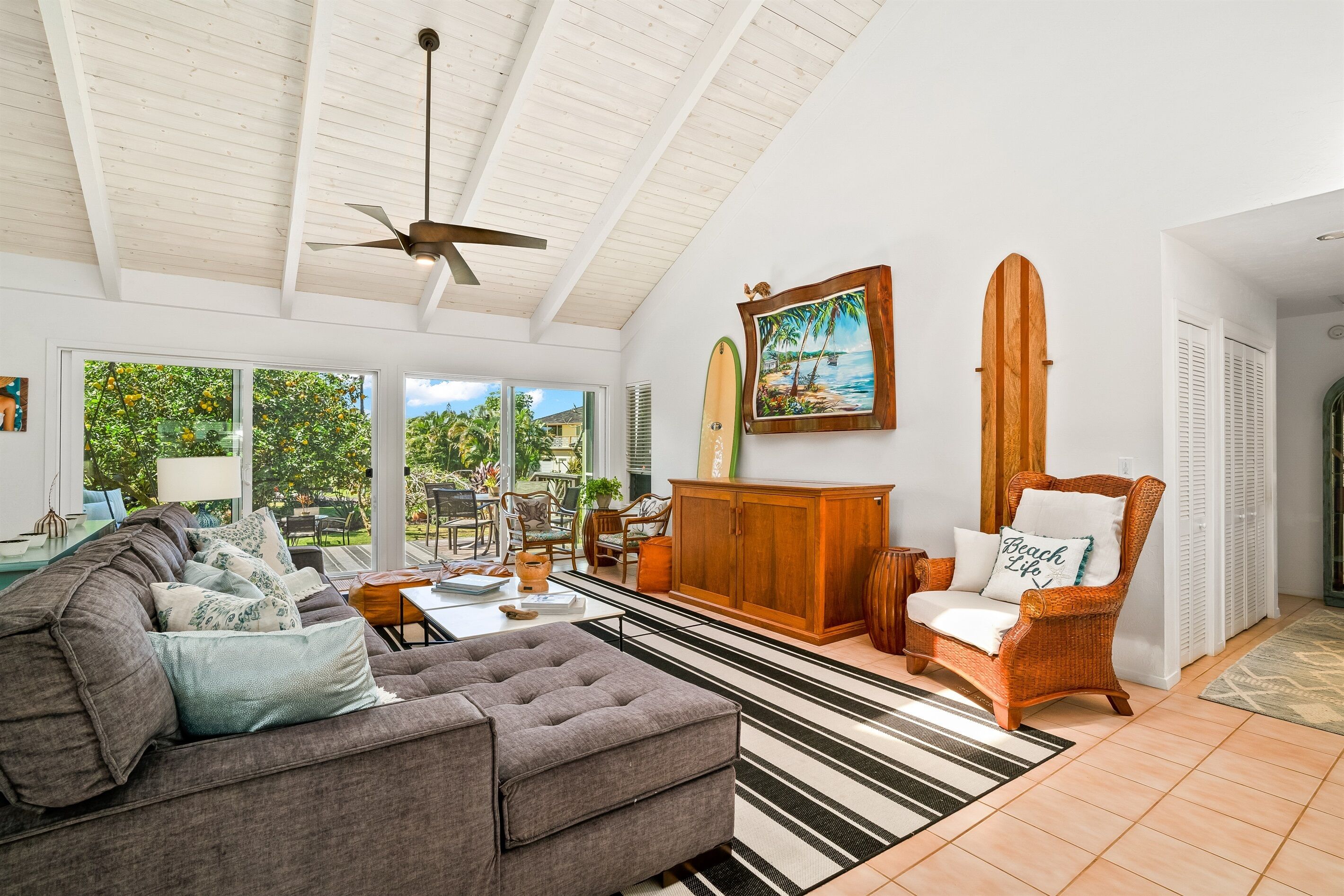 A spacious living room with a high ceiling, a ceiling fan, a large window with a view of greenery, a wooden cabinet, a wicker chair, and a couch with multiple cushions.