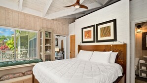 Une chambre avec un grand lit, une tête de lit en bois et un ventilateur de plafond. Une fenêtre offre une vue sur le mobilier et les plantes situés à l’extérieur.