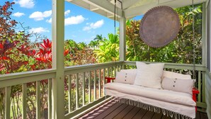 Une véranda avec une balançoire suspendue, un canapé blanc et une vue sur des plantes tropicales et un ciel dégagé.