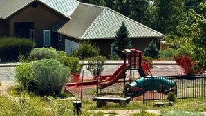 A building with a sloped roof, a playground with a slide, and a green vehicle parked nearby. - Modern Private Casita • Bright, Quiet, Minutes to LANL & Trails (Los Alamos)