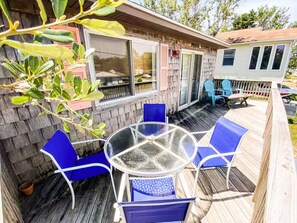 A covered outdoor patio with a round glass table and blue chairs, surrounded by wooden decking and a wooden fence. - Welcome to Sea Perch, a cozy beach box with a large hot tub! (Kill Devil Hills)