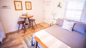 Ein Schlafzimmer mit Bett, Schreibtisch, zwei Stühlen, einem kleinen Tisch und einem Fenster mit Jalousien.