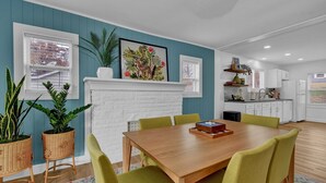 Un comedor con mesa de madera y sillas verdes, chimenea de ladrillo blanco y una cocina al fondo.