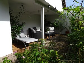 Un espacio de estar al aire libre cubierto, con un sofá de mimbre y una mesa, rodeado de plantas en macetas y una terraza de madera.
