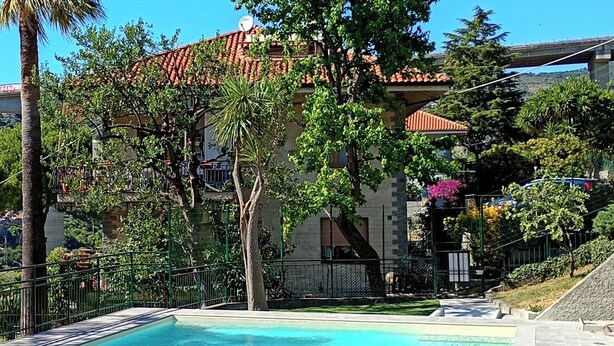 A swimming pool with a tiled deck, surrounded by greenery and a building with a red-tiled roof. - New two-room apartment with residential pool, large terrace and exclusive parking space (Bordighera)