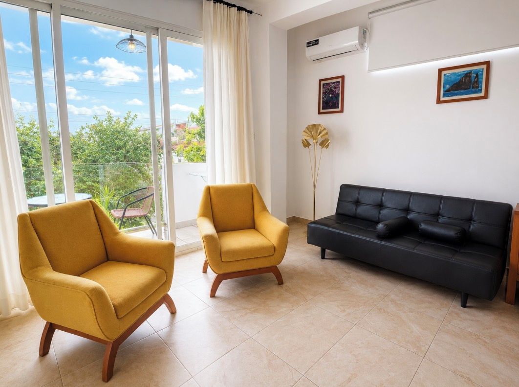 A living room with a sofa and two armchairs, a large window with curtains, and a view of greenery outside.