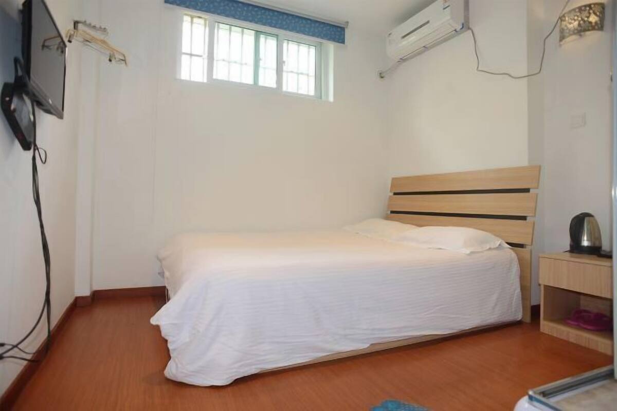 A single bed with a wooden headboard in a room with a window, an air conditioning unit, and a small bedside table with a lamp.