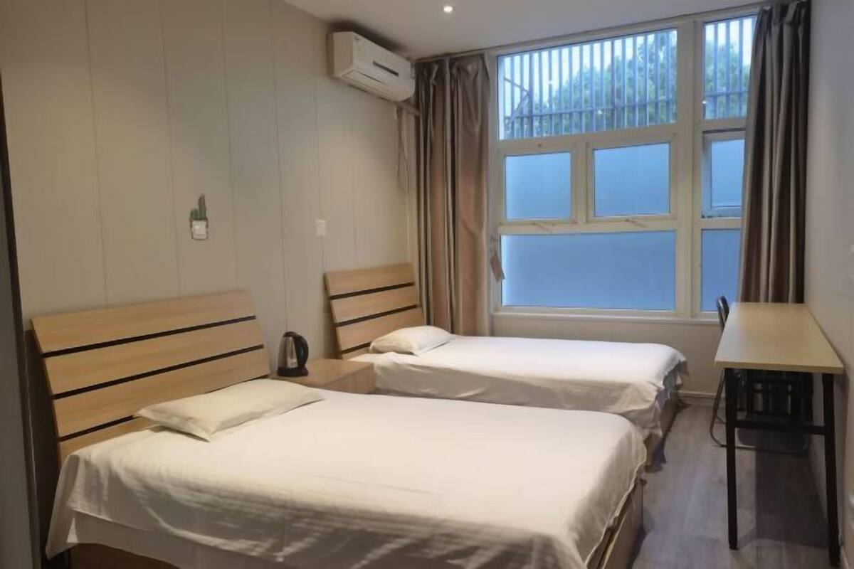 A room with two single beds, a wooden headboard, a small table, and a window with curtains.