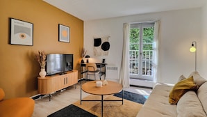 A modern living room with a television, sofa, and coffee table. There is a dining area with chairs and a table. The room has a large window with curtains. - Le Van Gogh - In the Heart of Arles (Arles)