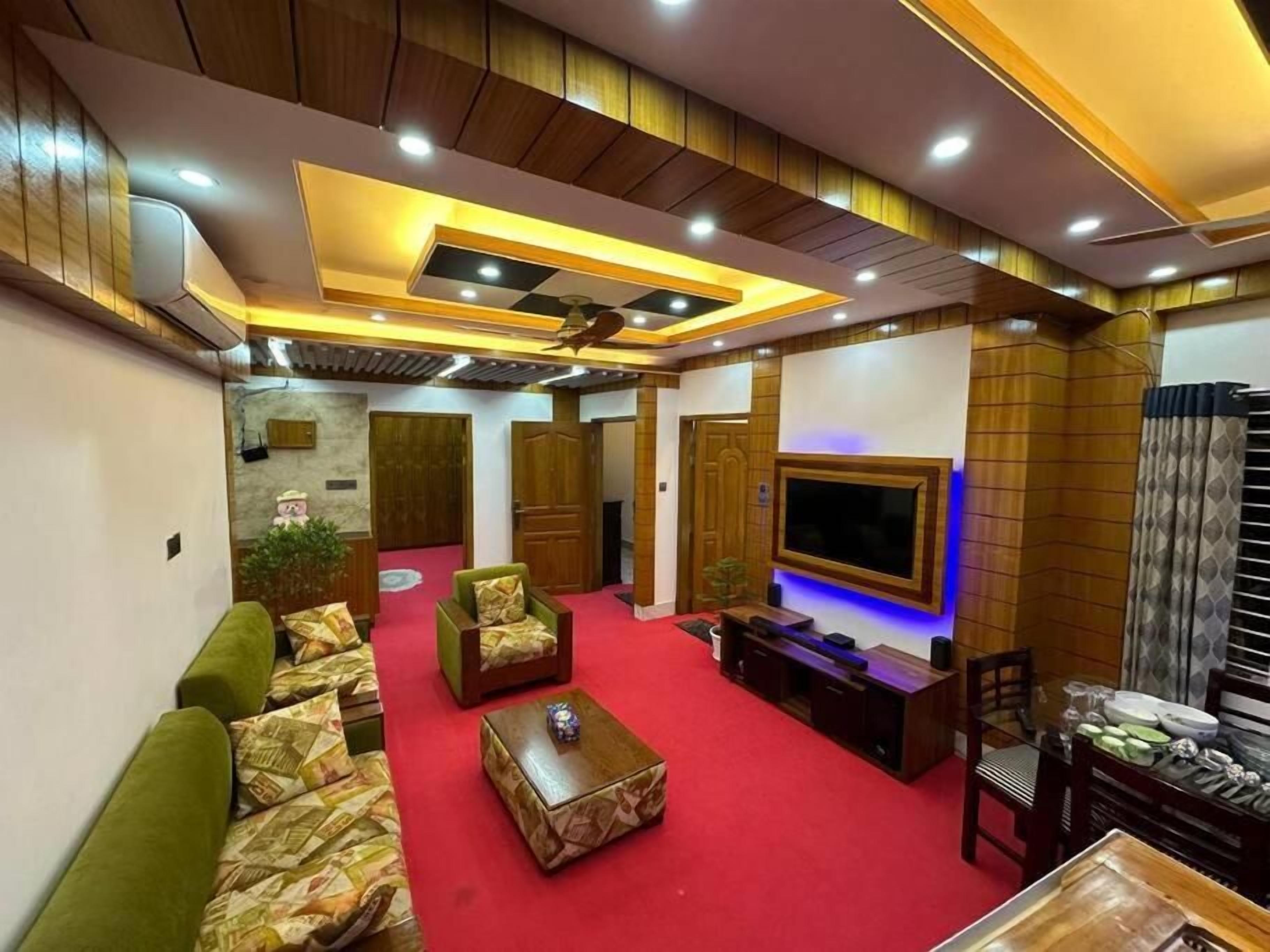 A spacious living room with a red carpet, green sofas, a coffee table, and a TV mounted on the wall.