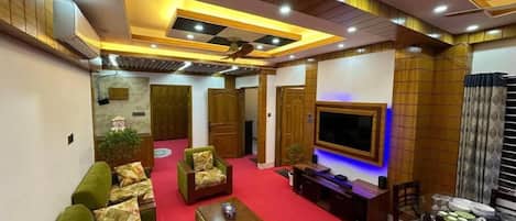 A spacious living room with a red carpet, green sofas, a coffee table, and a TV mounted on the wall.