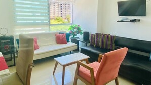 Una sala de estar con sofá, una silla, una mesa de centro, televisor montado en la pared y una ventana con persianas.