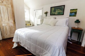 Un dormitorio con una cama grande, una puerta de madera, una ventana, una mesita de noche con una lámpara y un cuadro en la pared.