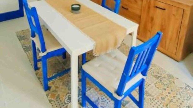 A compact kitchen with a wooden table and blue chairs, a hanging light fixture, and a TV mounted on the wall. - La Luna Guest House (Castellammare del Golfo)
