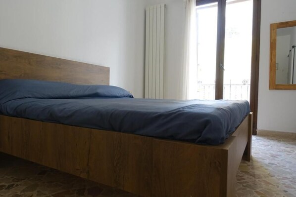 A single bed with a wooden headboard in a room with stone flooring and a window with curtains.