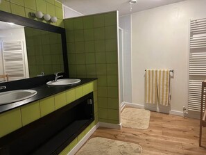 Kylpyhuoneessa on musta työtaso, kaksi pesuallasta, peili, vihreät laatat, suihku, pyyhekoukku ja puulattia.