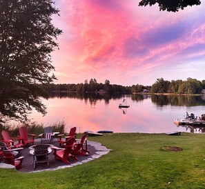 Una terraza junto al lago con sillas Adirondack rojas, un brasero y vistas al lago y árboles.