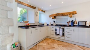 A kitchen with white cabinets, a wooden herringbone floor, a stone wall, and a window with a view of greenery.