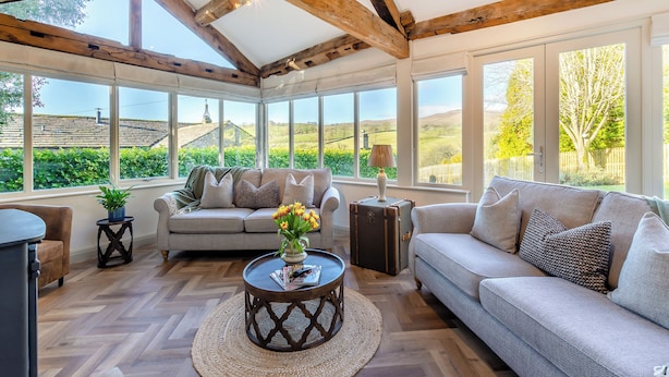 A conservatory with a wooden ceiling, large windows, a sofa, a coffee table, and a potted plant.