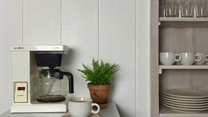 Una cafetera sobre una encimera con una taza y un plato, una planta en maceta y una estantería con copas y platos.