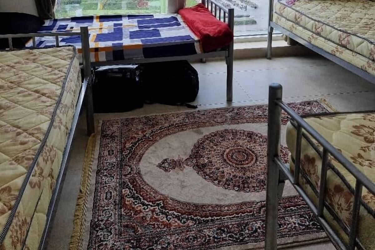 A dormitory room with two beds, a large window with curtains, a patterned rug, and a view of a cityscape and bridge.