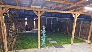 A covered patio area with a paved floor, a small grassy section, and a wooden fence. - NightRest Homes Hemel Hempstead 4 Bedroom 2 Toilet (Hemel Hempstead)
