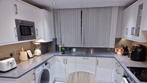 A compact kitchen with a microwave, toaster, washing machine, and a small dining table with a chair. - NightRest Homes Hemel Hempstead 4 Bedroom 2 Toilet (Hemel Hempstead)