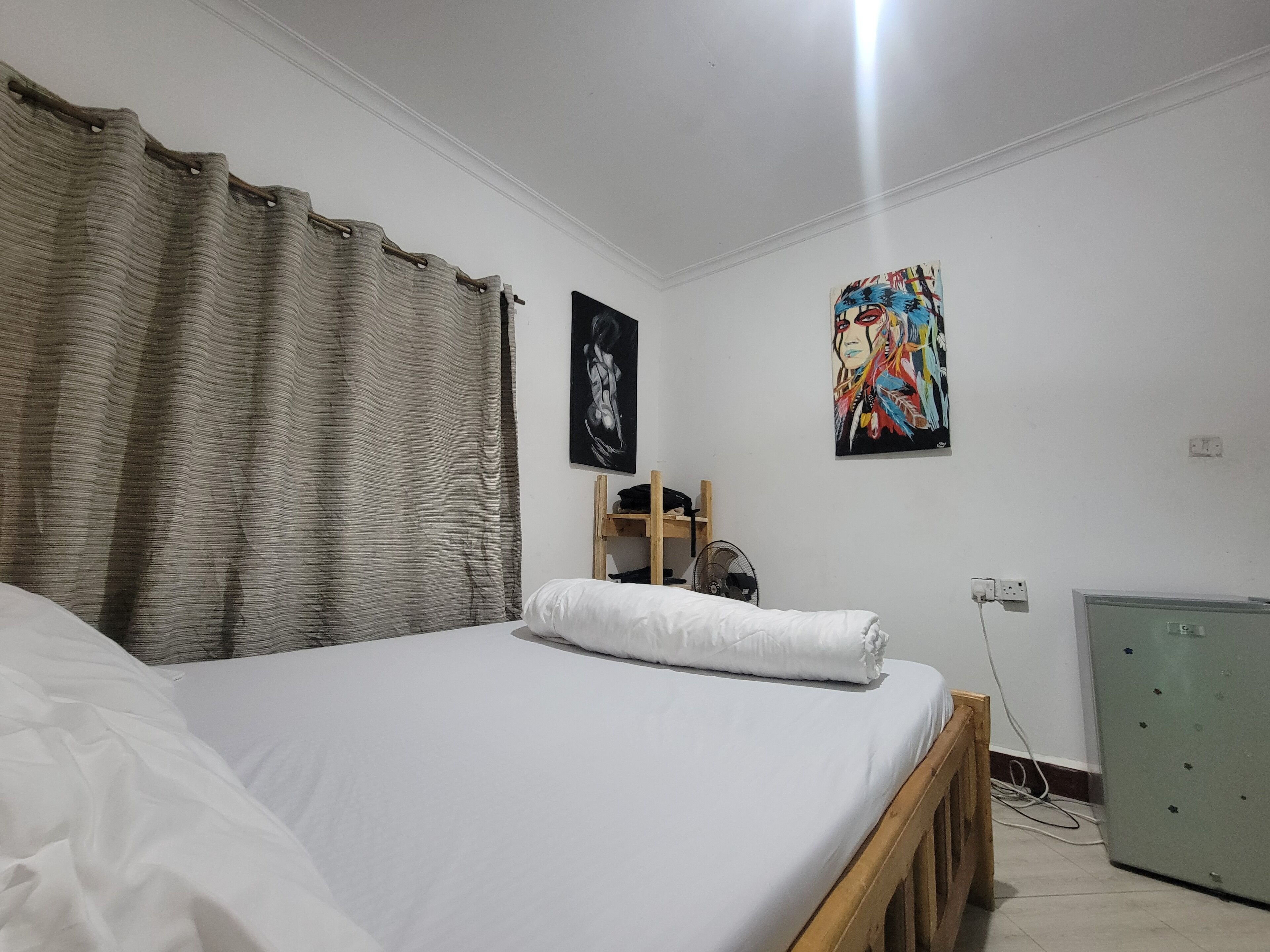 Une chambre à coucher avec un lit, une tête de lit en bois, un bureau avec une chaise, un ventilateur et une armoire verte.