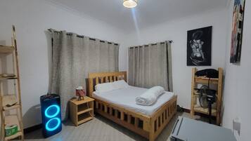 Quarto com cama de madeira, cortinas, ventilador, um alto-falante e um quadro na parede.