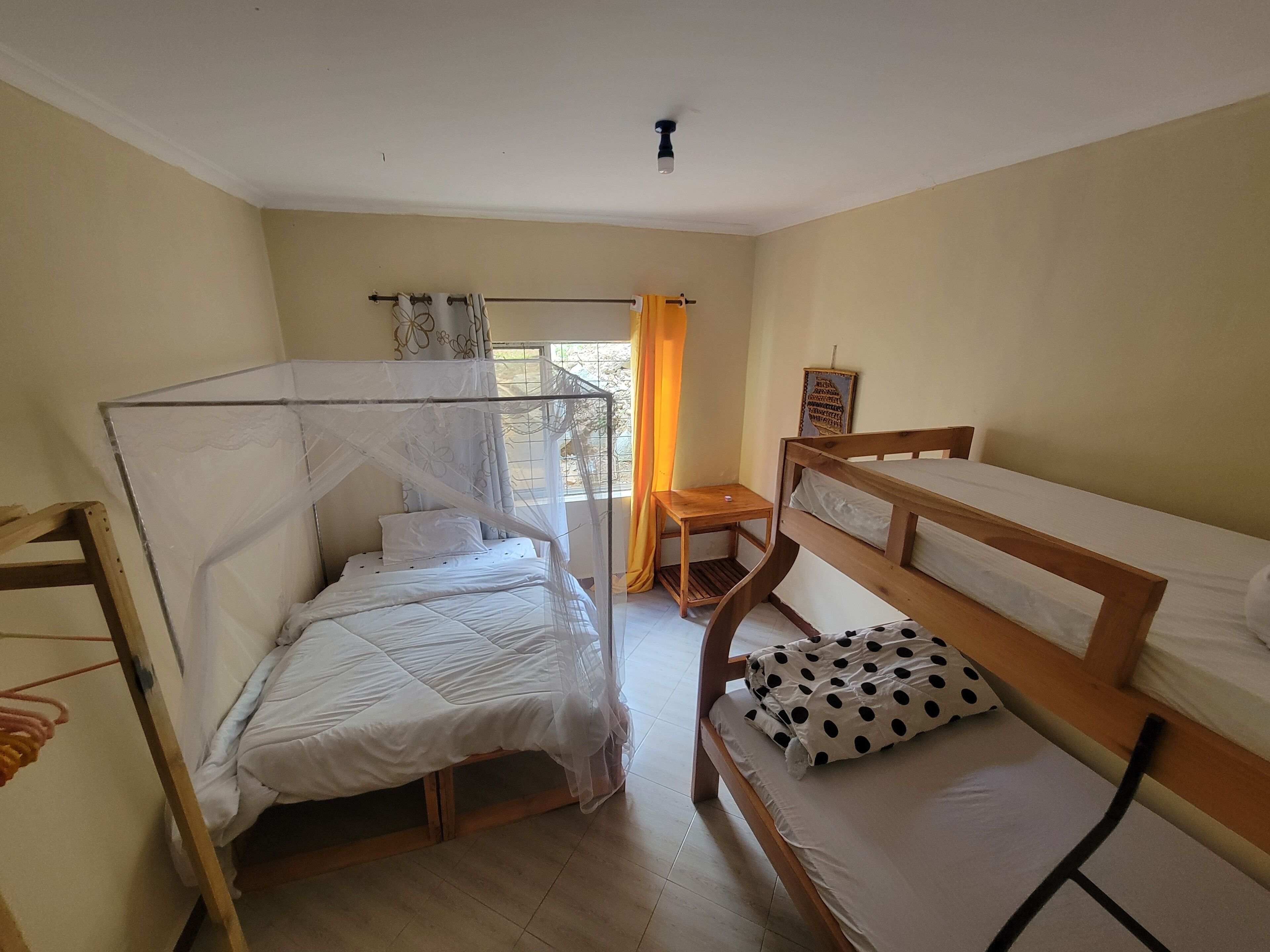 Ein Zimmer mit einem Bett, einer hölzernen Bank, einem kleinen Tisch und einem Fenster mit Vorhängen.